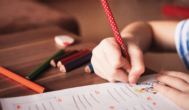 child's hand holding a pencil and marking on a page, coloured pencils scattered on the table