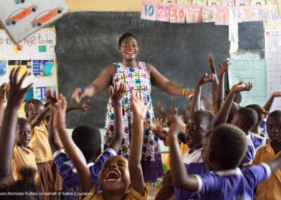 Teacher in primary classroom, smiling with her arms outstretched, the children are waving their hands in the air and shouting