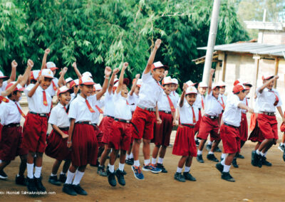 Asian children in school uniform - white shirt and dark red skirt/shorts jumping with hands raised outside a school