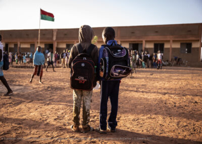 Two boys standing in the yard area of an African school, both wearing back packs. In the background is the school with other children assembling