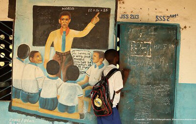 A student looking into his classroom, Sierra Leone, January 2015. On the wall by the door is a mural of a teacher and class with a child with a speech bubble - xxx is a very good maths teacher he makes us enjoy his lessons