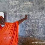 Female teacher ini a bright orange robe, stands at the blackboard in a classroom for the Kano Literacy and Mathematics Accelerator (KaLMA) project, Kano State, Nigeria.