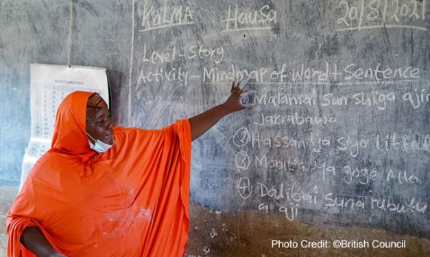 Female teacher ini a bright orange robe, stands at the blackboard in a classroom for the Kano Literacy and Mathematics Accelerator (KaLMA) project, Kano State, Nigeria.