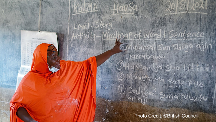 Female teacher ini a bright orange robe, stands at the blackboard in a classroom for the Kano Literacy and Mathematics Accelerator (KaLMA) project, Kano State, Nigeria.