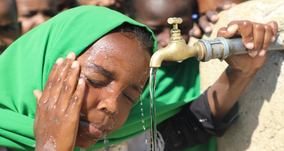 a young child splashes water on her face from the school tap, with other children waiting their turn behind her.