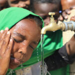 a young child splashes water on her face from the school tap, with other children waiting their turn behind her.