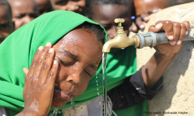 a young child splashes water on her face from the school tap, with other children waiting their turn behind her.