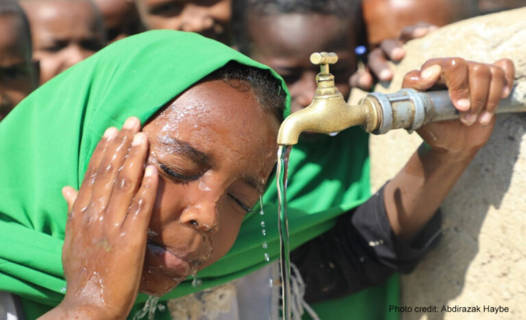 a young child splashes water on her face from the school tap, with other children waiting their turn behind her.
