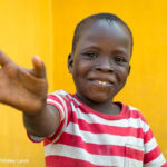 Young boy, against a yellow background, wearing a red and white striped shirt waving, Liberia, Sierra Leone.