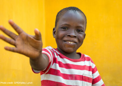 Young boy, against a yellow background, wearing a red and white striped shirt waving, Liberia, Sierra Leone.