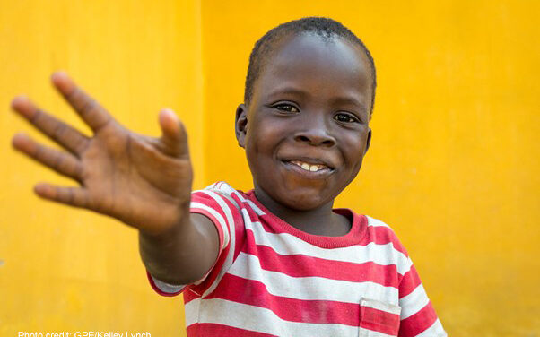 Young boy, against a yellow background, wearing a red and white striped shirt waving, Liberia, Sierra Leone.