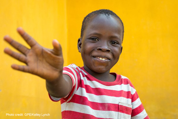 Young boy, against a yellow background, wearing a red and white striped shirt waving, Liberia, Sierra Leone.