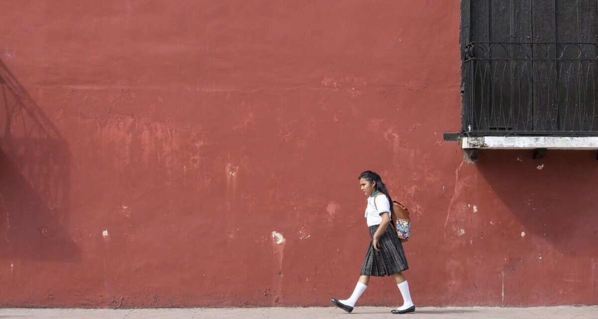 Girl walks to school in her uniform with a background of a dark red painted wall