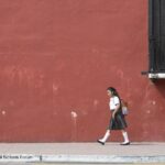 Girl walks to school in her uniform with a background of a dark red painted wall