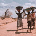 Two women and a girl carrying basins on their heads, while walking barefoot, Uganda.