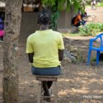 Adolescent girl in yellow t shirt sits alone outside a classroom, Uganda